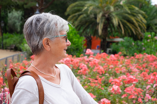 Smiling casual senior woman walking in the park looking at blooming flowers. Palm trees in background