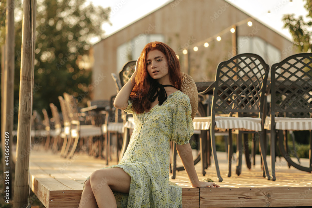Fashionable girl with red hair and freckles in summer stylish dress and black bandage on neck looking into camera, relaxing in country cafe