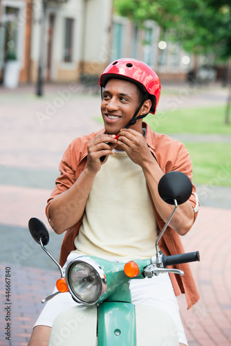 smiling african american man on scooter fastening protective helmet