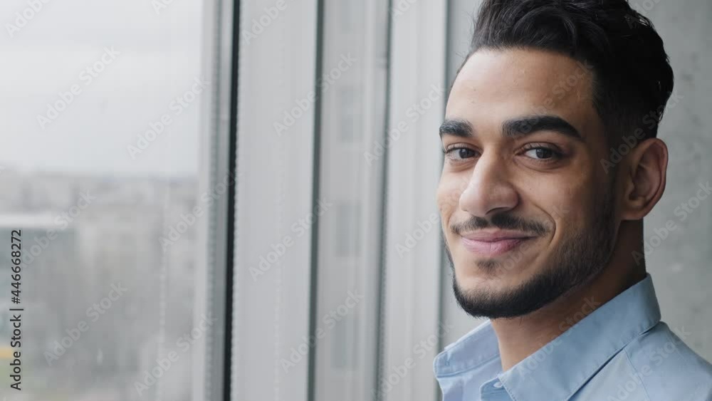 Side view of young adult handsome arabic man hispanic male boss leader employer successful professional specialist businessman stands near window in modern company turns head looks at camera smiling Side view of young adult handsome arabic man hispanic male boss leader employer successful professional specialist businessman stands near window in modern company turns head looks at camera smiling