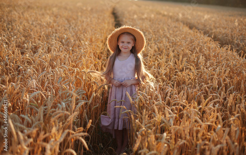 a beautiful blonde girl with long hair in a pink linen dress and a straw hat stands in a wheat field in the setting sun © Тамара Киреева