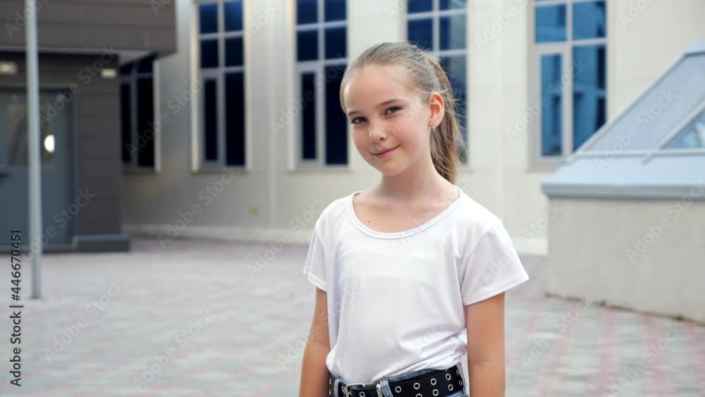 Girl model poses looking straight. Long haired blonde teenage girl in white t-shirt stands near school building smiling on sunny summer day close view Girl model poses looking straight. Long haired blonde teenage girl in white t-shirt stands near school building smiling on sunny summer day close view