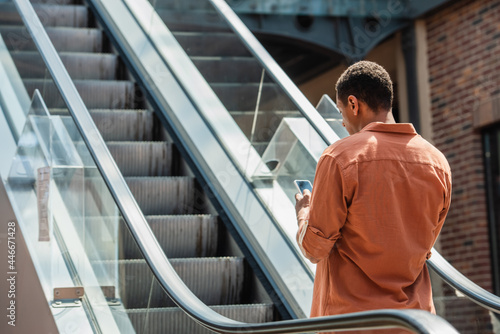young african american man in shirt messaging on mobile phone on escalator