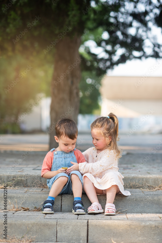 A cute boy and a curly girl are sitting on a step under a tree in the park and looking at something A cute boy and a curly girl are sitting on a step under a tree in the park and looking at something