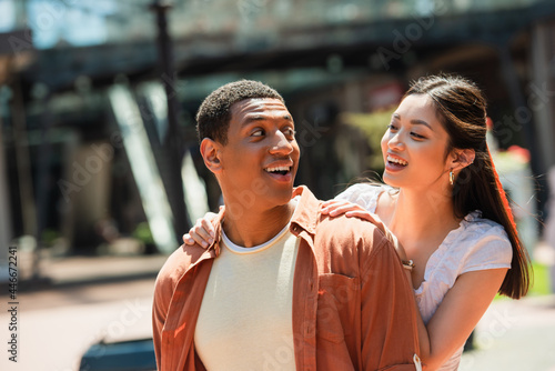 excited multiethnic couple smiling at each other while walking in city