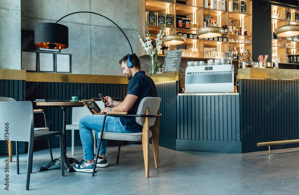 Casual Businessman with Headphones Working on Laptop and Mobile Phone in a Cafe.
Young business man using phone while working on laptop and drinking coffee in a restaurant.