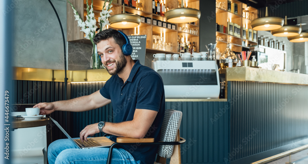 Cheerful Businessman Looking at Camera while Working on Laptop in a Cafe.
Portrait of smiling business man teleconferencing using laptop computer and headphones while drinking coffee in a restaurant.