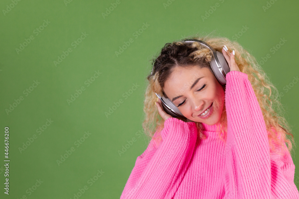 Young blonde woman with long curly hair in pink sweater on green background happy positive enjoying favorite music in wireless headphones Young blonde woman with long curly hair in pink sweater on green background happy positive enjoying favorite music in wireless headphones