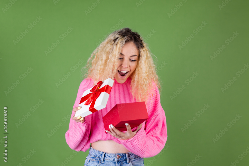 Young blonde woman with long curly hair in pink sweater on green background with gift box happy cheerful excited smiling Young blonde woman with long curly hair in pink sweater on green background with gift box happy cheerful excited smiling