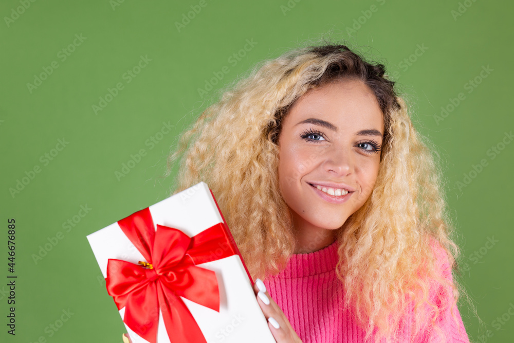 Young blonde woman with long curly hair in pink sweater on green background with gift box happy cheerful excited smiling Young blonde woman with long curly hair in pink sweater on green background with gift box happy cheerful excited smiling