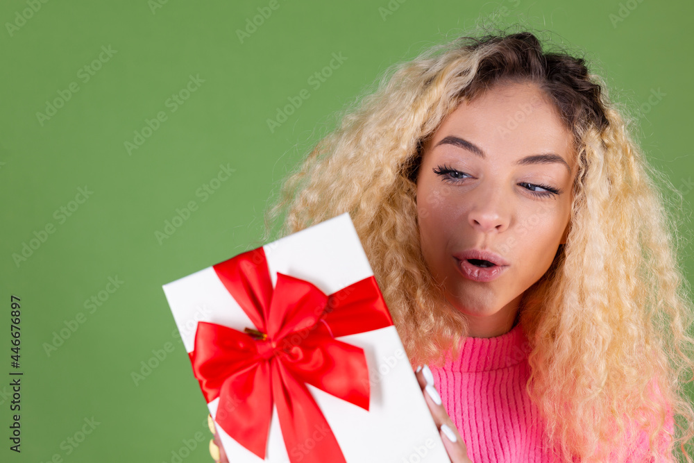 Young blonde woman with long curly hair in pink sweater on green background with gift box happy cheerful excited smiling Young blonde woman with long curly hair in pink sweater on green background with gift box happy cheerful excited smiling