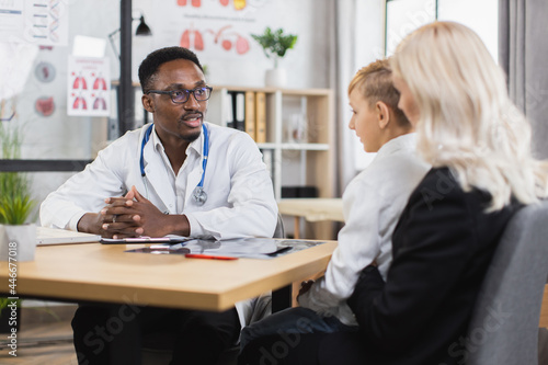 African american pediatrician in white lab coat sitting at his cabinet and providing medical consultation for charming woman with son. Caucasian mother with child having regular visit to doctor.