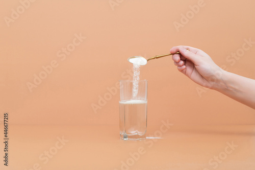 Hand pours collagen protein powder in a glass of water on a beige background. A natural supplement for skin beauty and bone health.