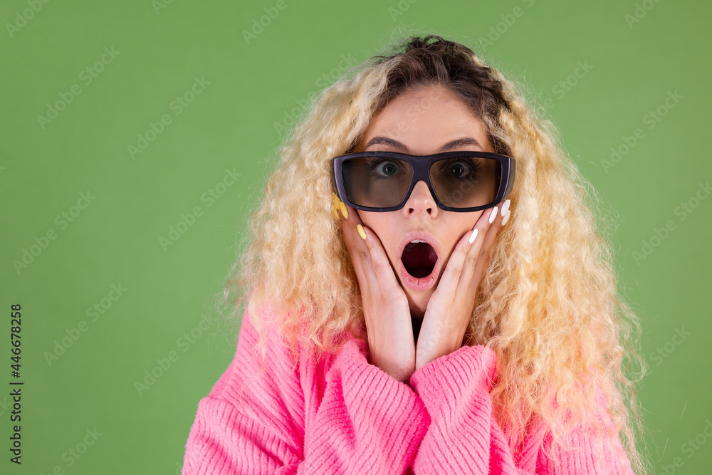 Young blonde woman with long curly hair in pink sweater on green background in 3d cinema shocked Young blonde woman with long curly hair in pink sweater on green background in 3d cinema shocked