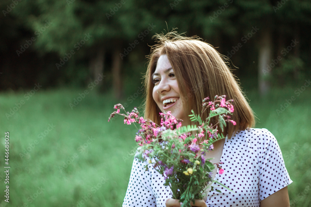 Fototapeta premium A girl with a short haircut and flowers in her hands looks away and laughs. A close portrait of a young beautiful woman in a meadow with lilac wildflowers in her hands. Selective focus. Copy space
