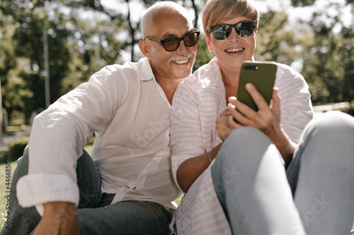 Joyful lady with short hair in sunglasses and striped shirt smiling, holding smartphone and opposing with happy old man with mustache in park.. © Look!