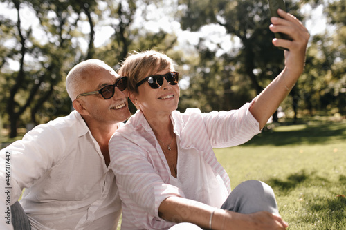 Beautiful lady with short hair in sunglasses, pink blouse and jeans sitting on grass and making photo with grey haired man in white outfit on park.. © Look!