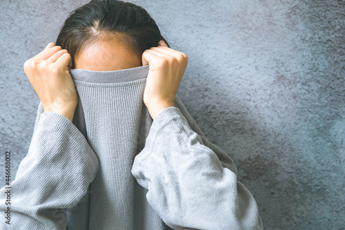 Asian woman hiding face under the clothes. She is oulling sweater on her head. She has social phobia, tying to be anonym.