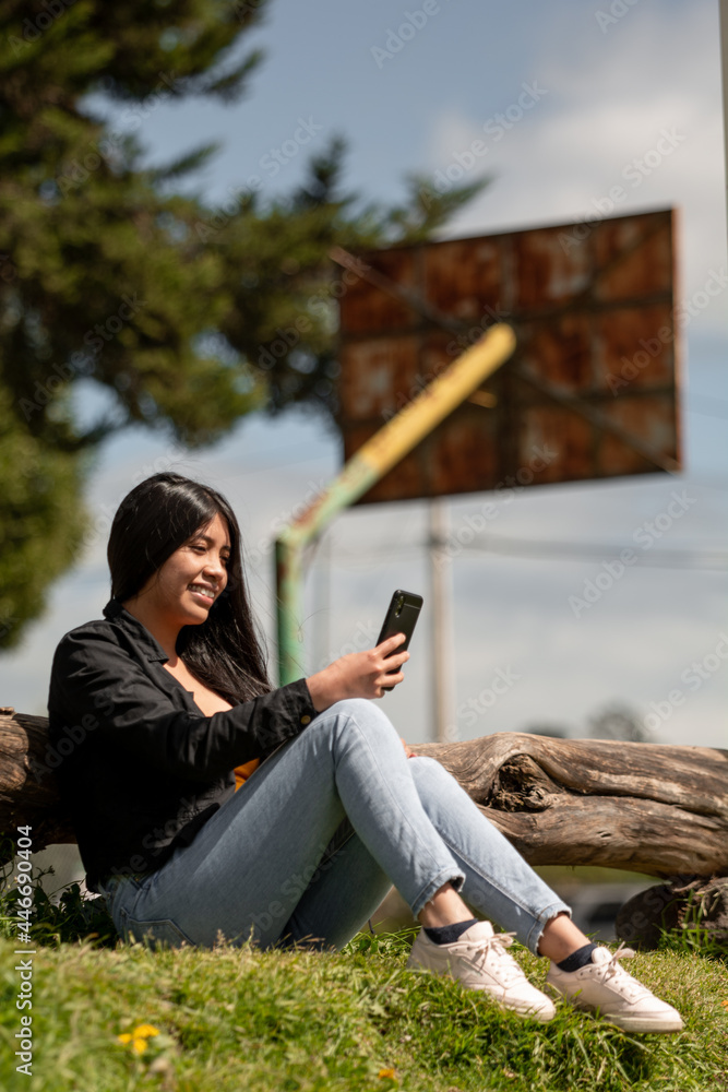 happy young latina woman with long hair sitting on the grass using her cell phone