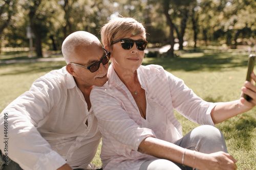 Blonde lady in sunglasses, striped stylish blouse and jeans. sitting on grass and making selfie with mustachioed man in white shirt outdoor.. © Look!