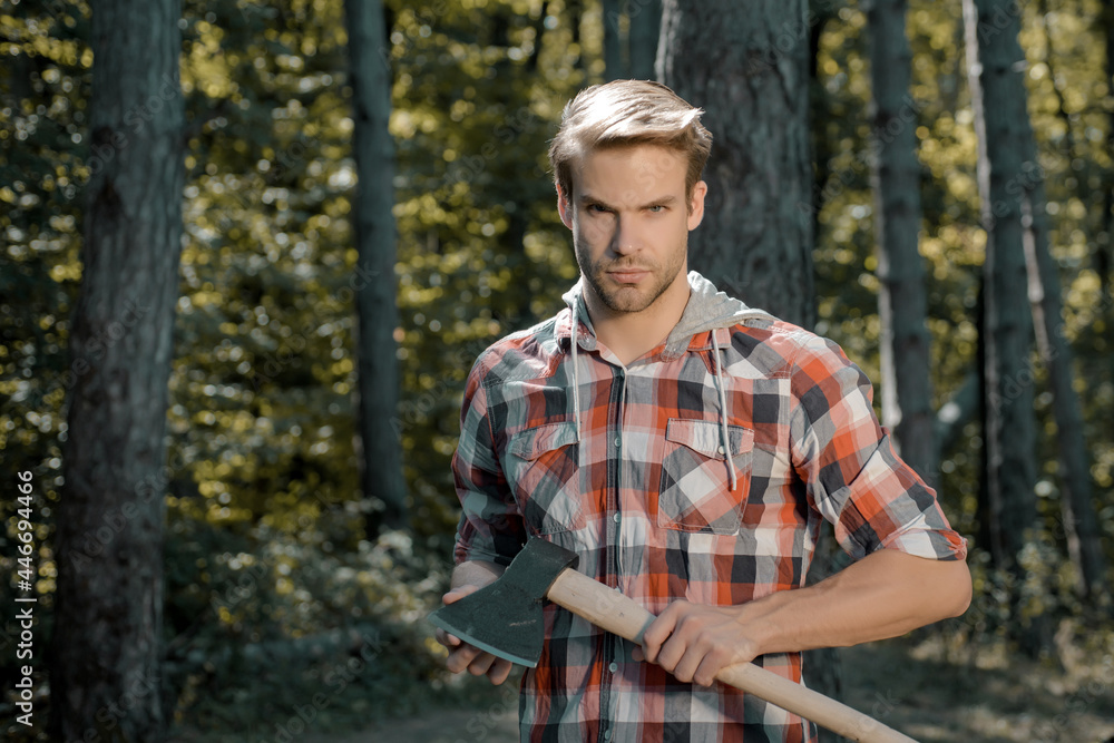 Firewood as a renewable energy source. Handsome Woodworkers lumberjack plaid shirt holding the axe on green nature background. Lumberjack worker standing in the forest with axe. Firewood as a renewable energy source. Handsome Woodworkers lumberjack plaid shirt holding the axe on green nature background. Lumberjack worker standing in the forest with axe.