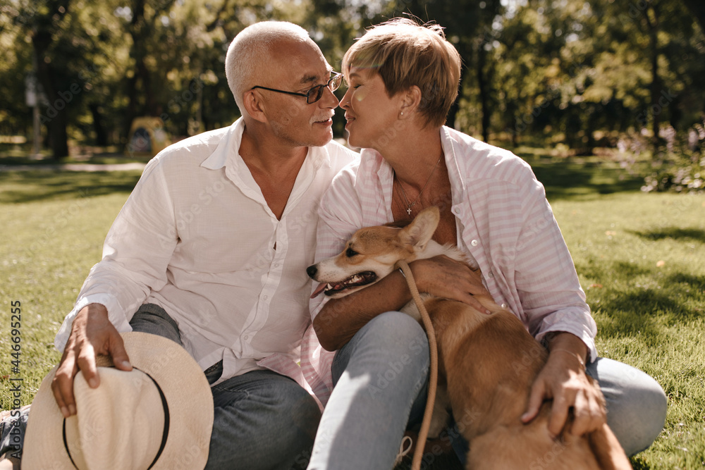 Short haired in striped pink blouse posing with small corgi and kissing man with mustache in white shirt and jeans in park.. Short haired in striped pink blouse posing with small corgi and kissing man with mustache in white shirt and jeans in park..