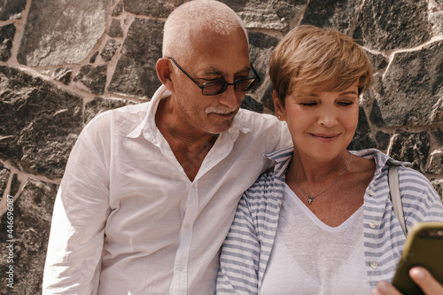 Old man with grey hair and mustache in light shirt and glasses looking at phone with blonde lady in striped outfit on backdrop of wall.. © Look!