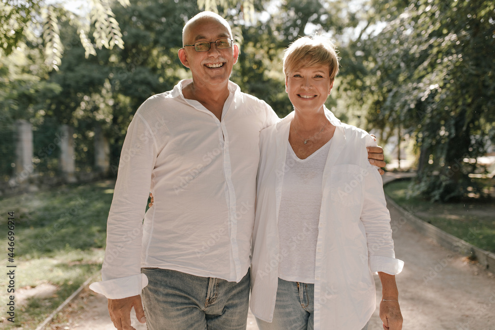 Old man with grey mustache and eyeglasses in white stylish shirt and jeans hugging her smiling wife with blonde hair in light blouse in park.. Old man with grey mustache and eyeglasses in white stylish shirt and jeans hugging her smiling wife with blonde hair in light blouse in park..