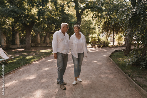Full length photo of happy man with grey hair in white shirt and jeans smiling and posing with blonde lady in light blouse in park.. © Look!