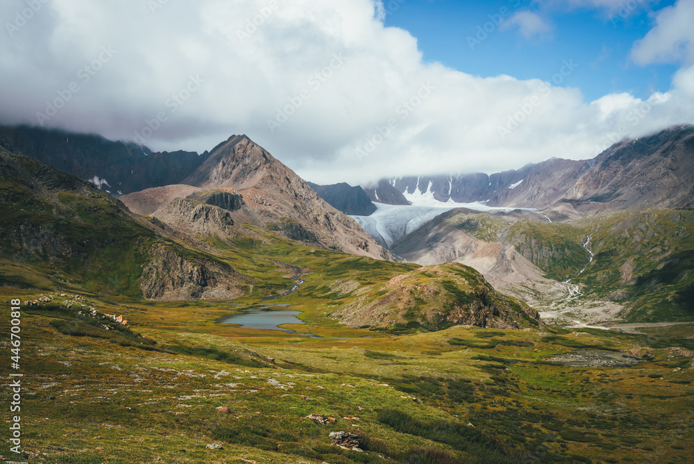 Fototapeta premium Scenic alpine landscape with mountain lake in sunlit green valley and glacier under cloudy sky. Awesome sunny scenery with beautiful glacial lake in sunlight against great mountain range in low clouds