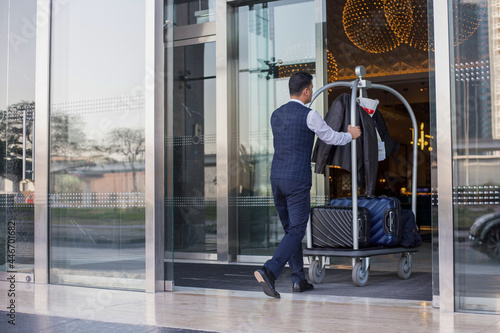 Young man pushes luggage cart with suitcases, bags and backpacks to the entrance. Metall baggage trailer with luggage