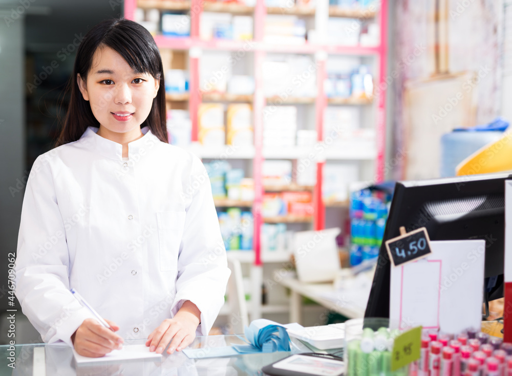 Smiling chinese female pharmacist standing with a cash desk in the pharmacy
