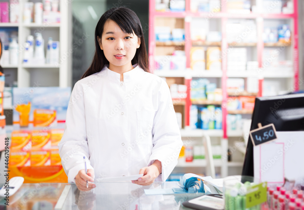 Cheerful chinese female pharmacist standing with a cash desk in the pharmacy