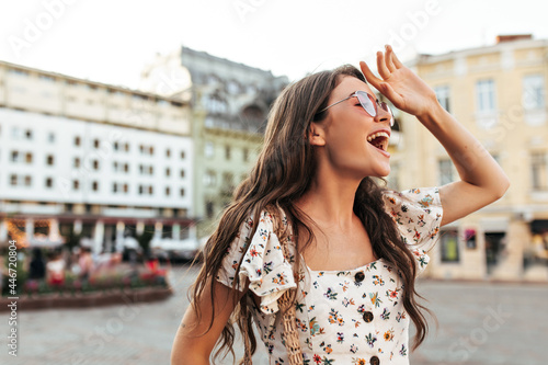 Charming brunette woman in stylish white floral blouse and trendy sunglasses smiles widely and looks away outdoor.
