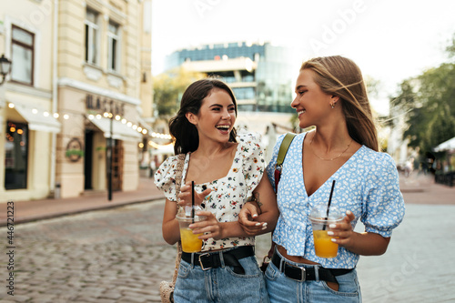 Beautiful charming brunette and blonde tanned girls in trendy colorful floral blouses talks  laugh  walk outside and holds glasses of lemonade.