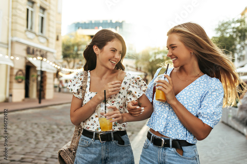 Cute young brunette and blonde women in stylish colorful blouses and denim pants look at each other, smile, walk outside and enjoy lemonade.