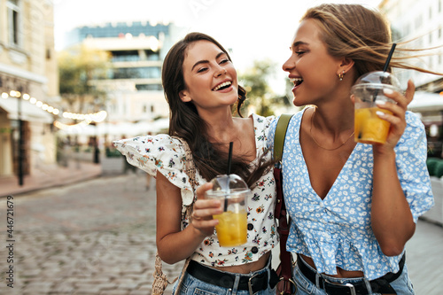 Happy young tanned brunette and blonde women in stylish cropped floral blouses smile sincerely  talk  walk in city center and enjoy fresh lemonade.