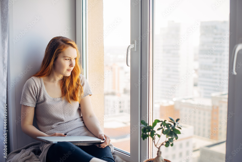 Relaxed long-haired brunette sitting on the windowsill, resting from work on laptop, thinking and looking at the big city outside the window. Freelance, self-isolation concept. Relaxed long-haired brunette sitting on the windowsill, resting from work on laptop, thinking and looking at the big city outside the window. Freelance, self-isolation concept.