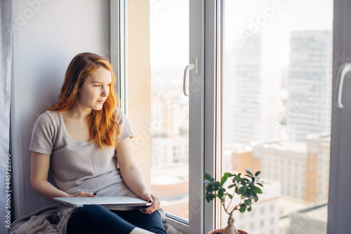 Relaxed long-haired brunette sitting on the windowsill, resting from work on laptop, thinking and looking at the big city outside the window. Freelance, self-isolation concept. Free Download Relaxed long-haired brunette sitting on the windowsill, resting from work on laptop, thinking and looking at the big city outside the window. Freelance, self-isolation concept. - GFXLoad