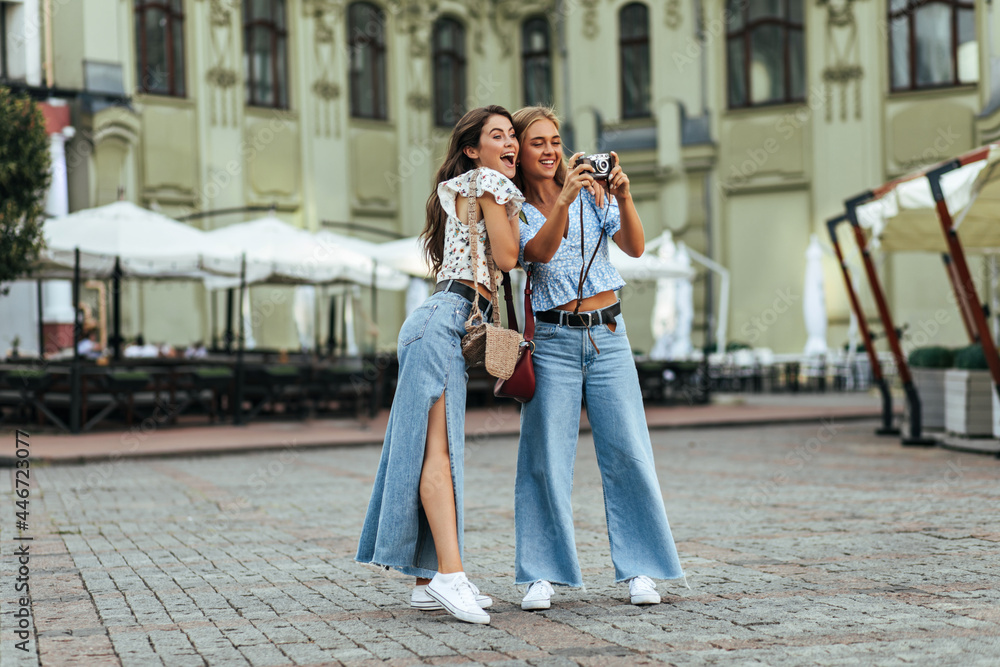 Surprised brunette woman in stylish denim pants and white floral blouse poses with her friend outside. Blonde lady in jeans and blue top takes photos on retro camera.