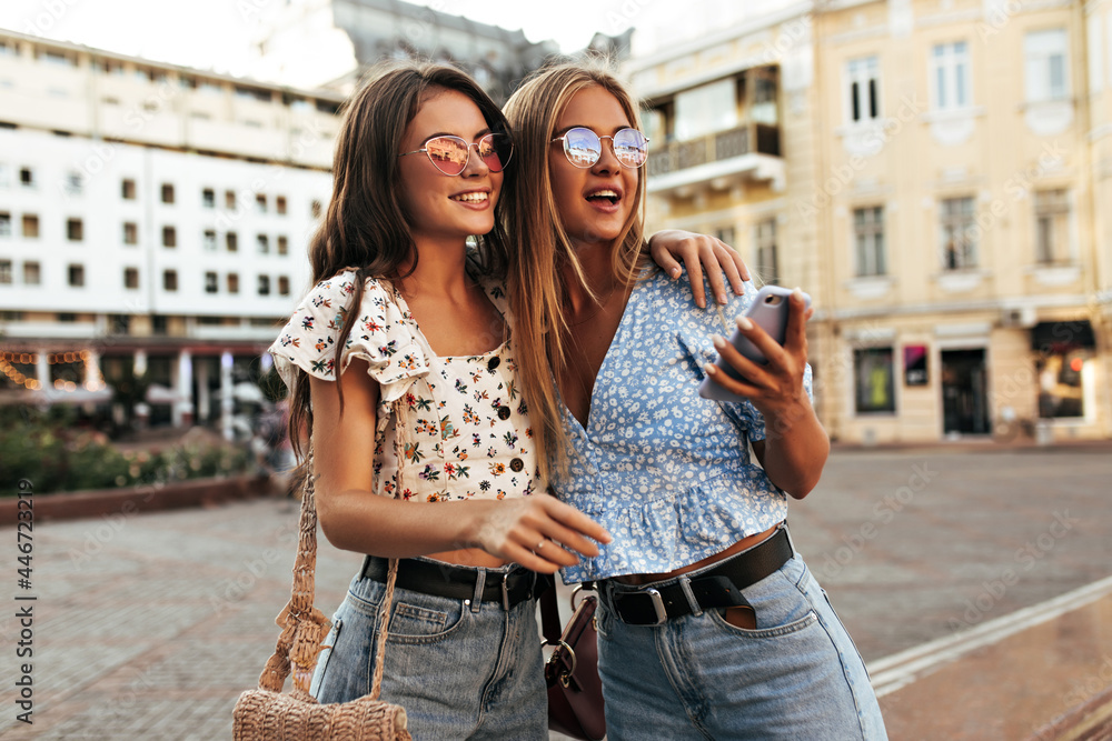 Attractive tanned girls in loose stylish jeans and floral blouses smile and hug. Blonde and brunette women pose outdoors. Charming lady holds phone.