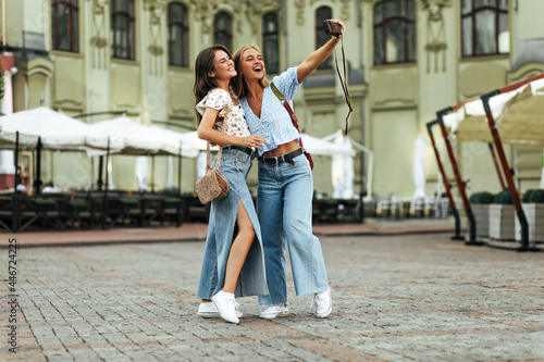 Brunette and blonde women in jeans and floral blouses take selfie outside. Happy young lady in denim pants and blue top holds retro camera and hugs friend.