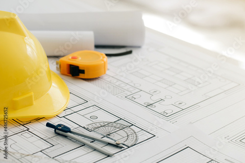 Yellow hard safety helmet hat and the blueprint, pen, ruler, protractor, and tape measure on the table at the construction site.for safety project of workman as engineer or worker, 