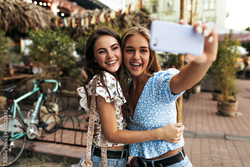 Joyful brunette and blonde women hug and take selfie outdoors. Young attractive girl in blue blouse holds cellphone and smiles.