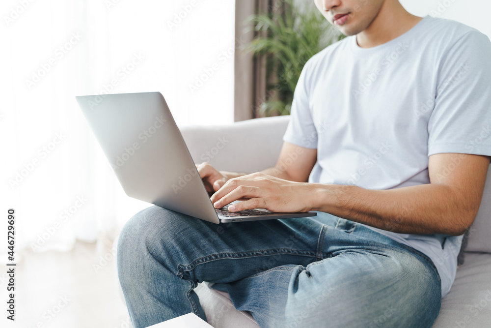 Naklejka premium Young man sitting on the sofa at home typing on the laptop keyboard for online working, using internet, freelancer, relaxed, modern job lifestyle, work at home..