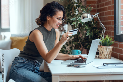 Cute woman holding white credit card for shopping online with computer while sitting in living room at home.