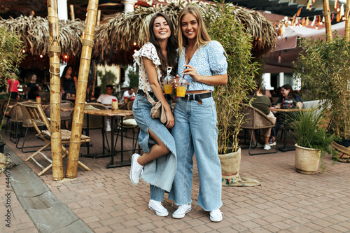 Cheerful curly brunette woman in trendy denim pants and white blouse walks with beautiful friend. Attractive blonde lady holds glass of lemonade.
