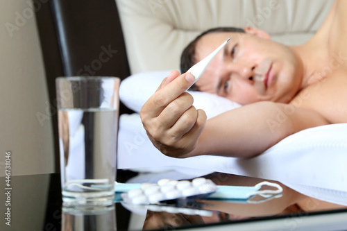 Sick man lying with digital thermometer in a bed  pills and water glass in the foreground. Concept of illness  fever  coronavirus symptoms