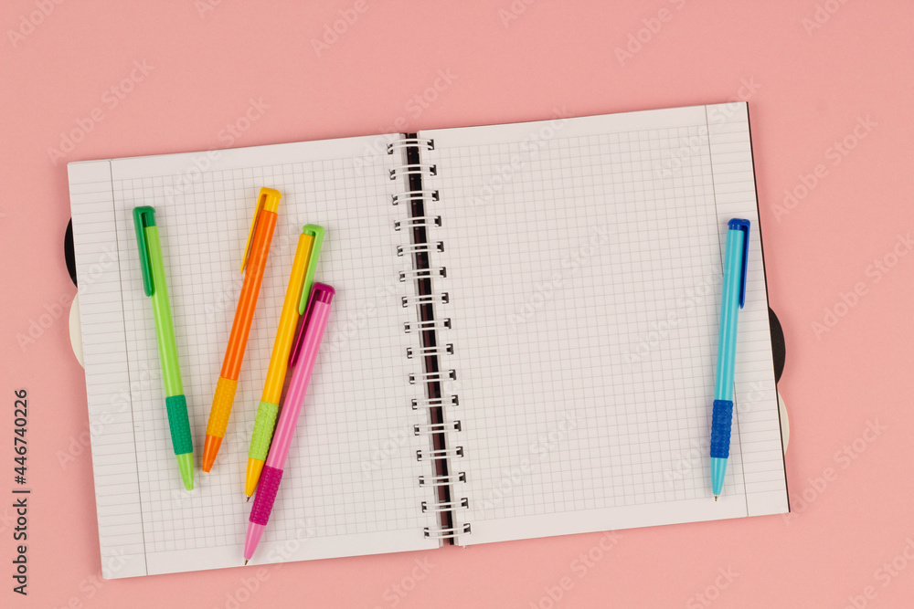 Open school squared notebook and colorful pens on the pink background. Blank white sheet of paper book on the table. Office supplies on the desktop. Back to school. Copy space for text. Top view Open school squared notebook and colorful pens on the pink background. Blank white sheet of paper book on the table. Office supplies on the desktop. Back to school. Copy space for text. Top view