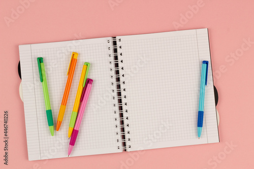Open school squared notebook and colorful pens on the pink background. Blank white sheet of paper book on the table. Office supplies on the desktop. Back to school. Copy space for text. Top view